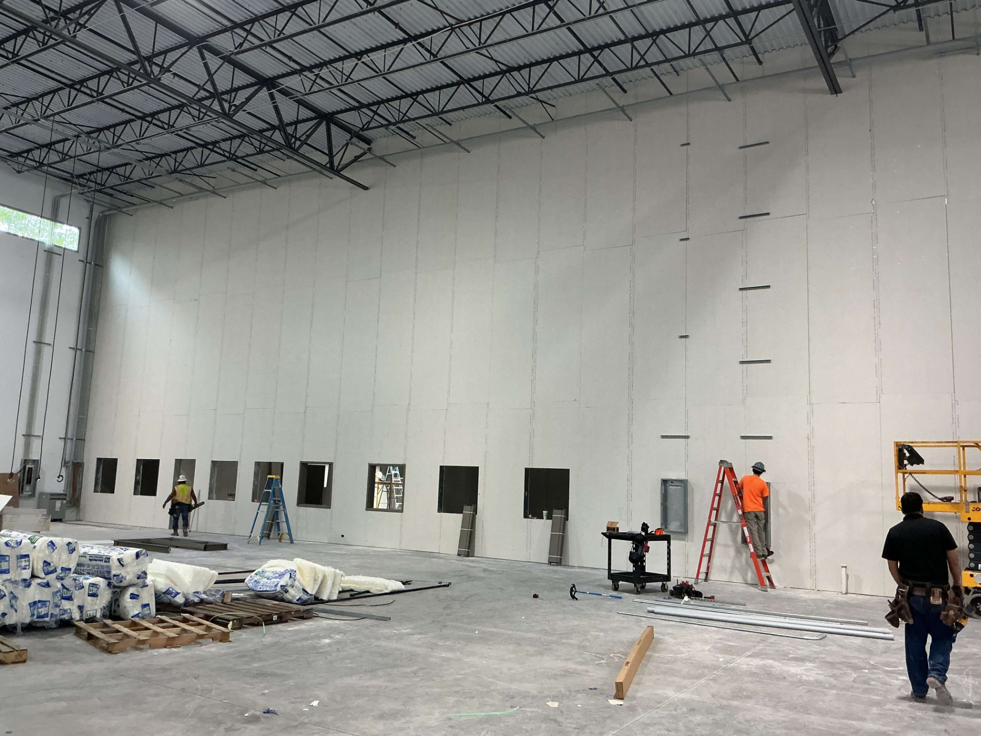 Large commercial interior with drywall hung on tall walls, workers on ladders and lifts